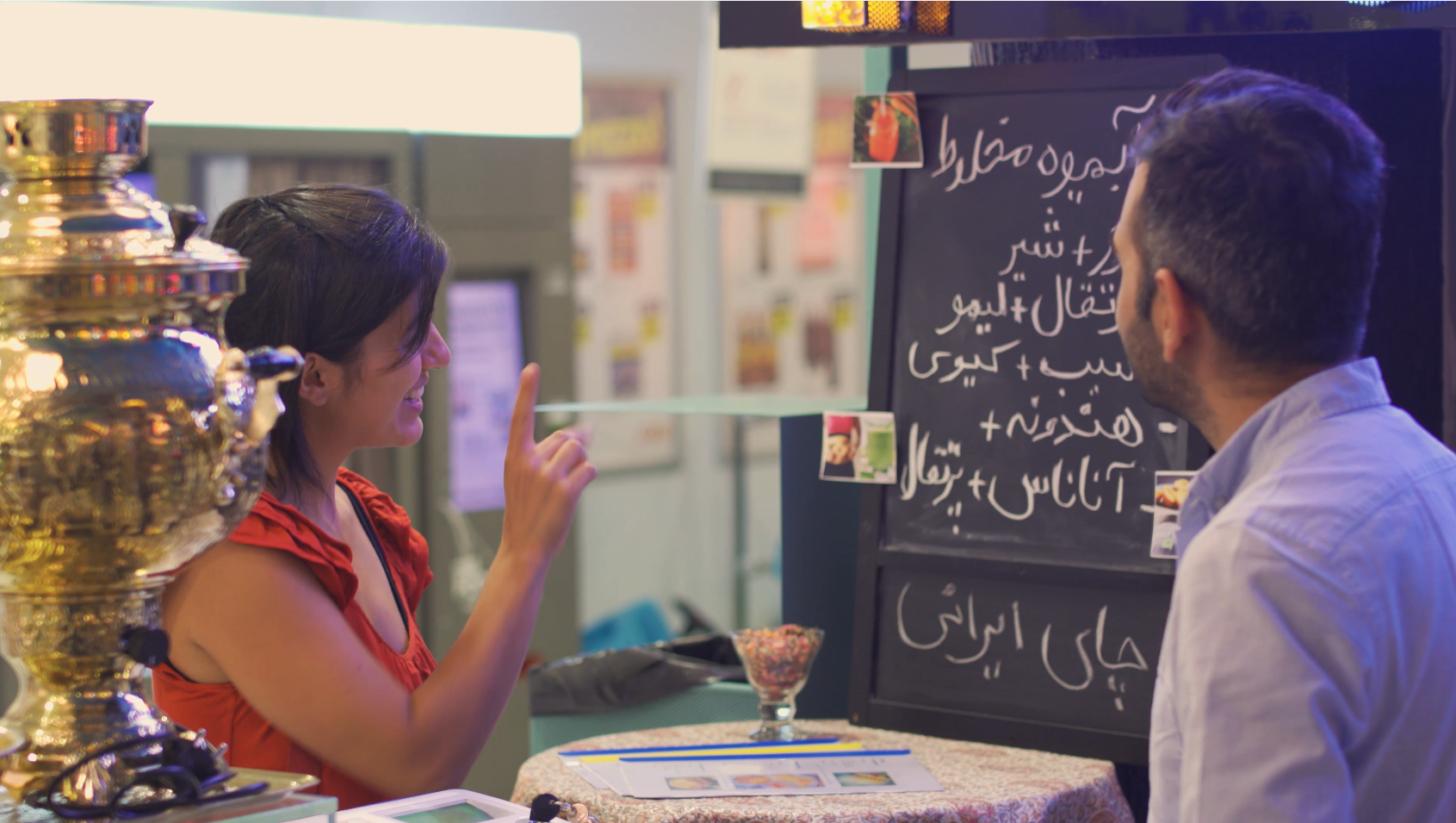 Una ragazza indica una lavagnetta con scritte in arabo al bancone di un bar, il barista la ascolta