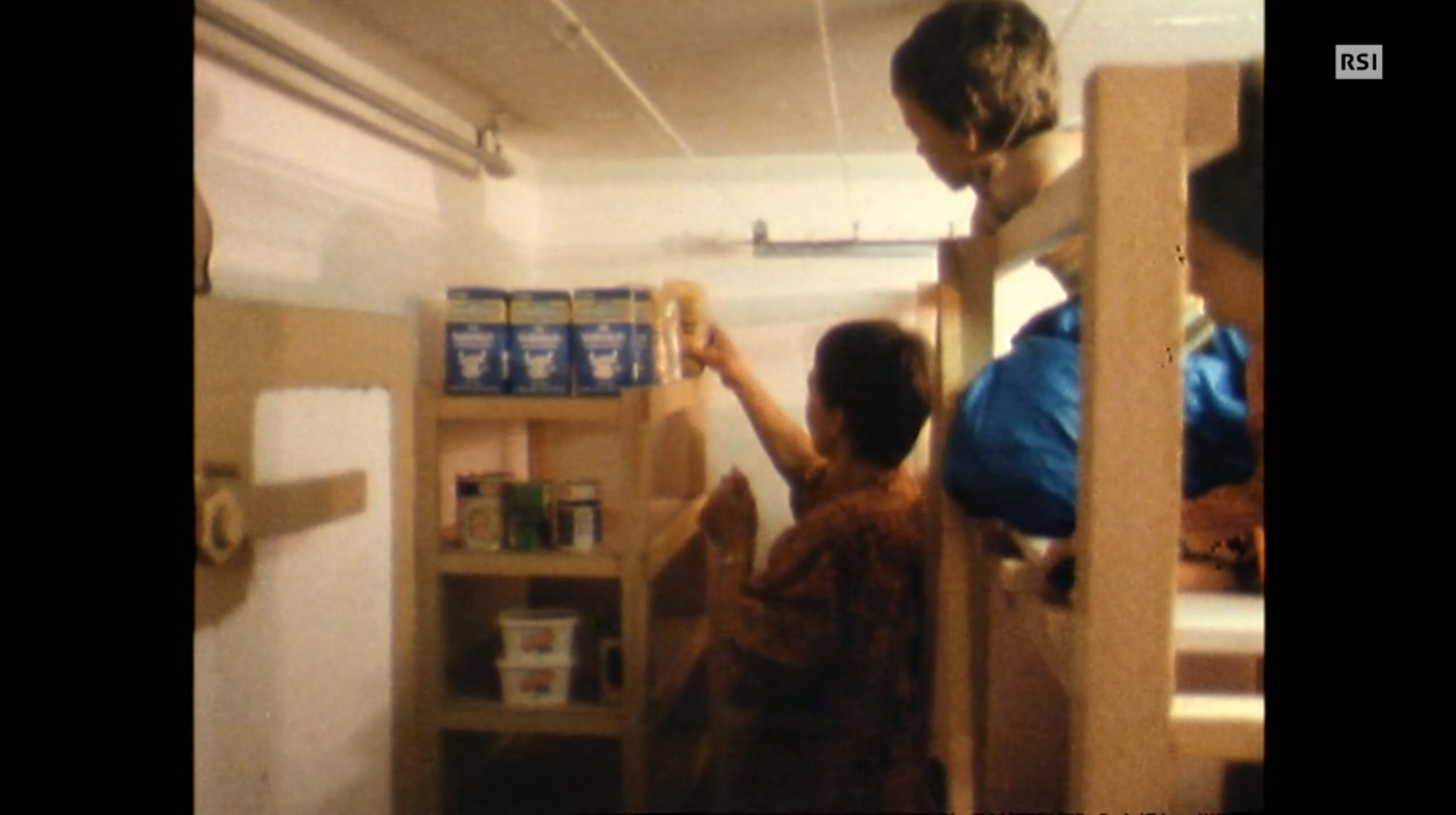 Interno d un bunker (porta in cemento armato sulla sx): signora sistema del cibo su degli scaffali accanto a un letto a castello