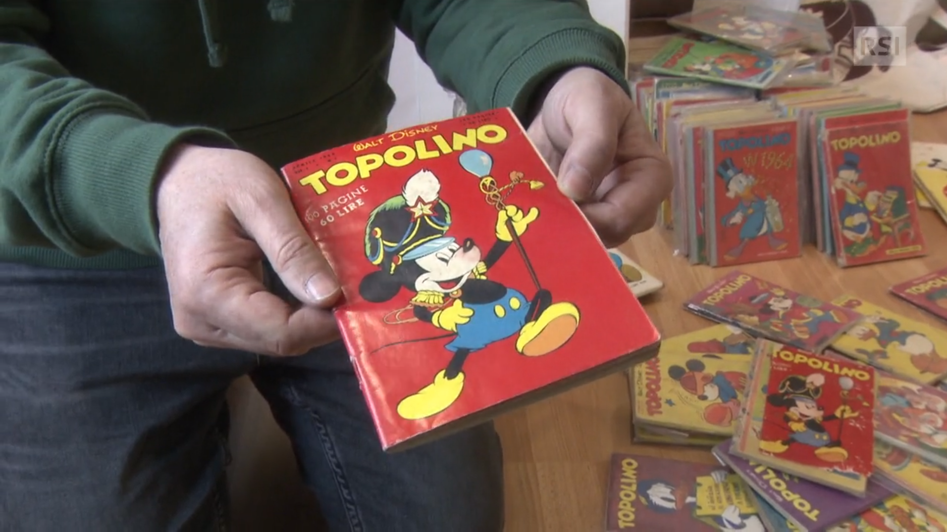 Primo piano di un numero di Topolino (l 1 in italiano) retto tra le mani da un uomo con maglione verde; sullo sfondo altri fumet