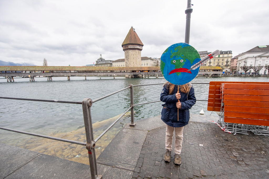 Eine junge Frau steht an einem Fluss in einer Stadt, mit einem runden Plakat, das die Erde als kranken Patienten zeigt