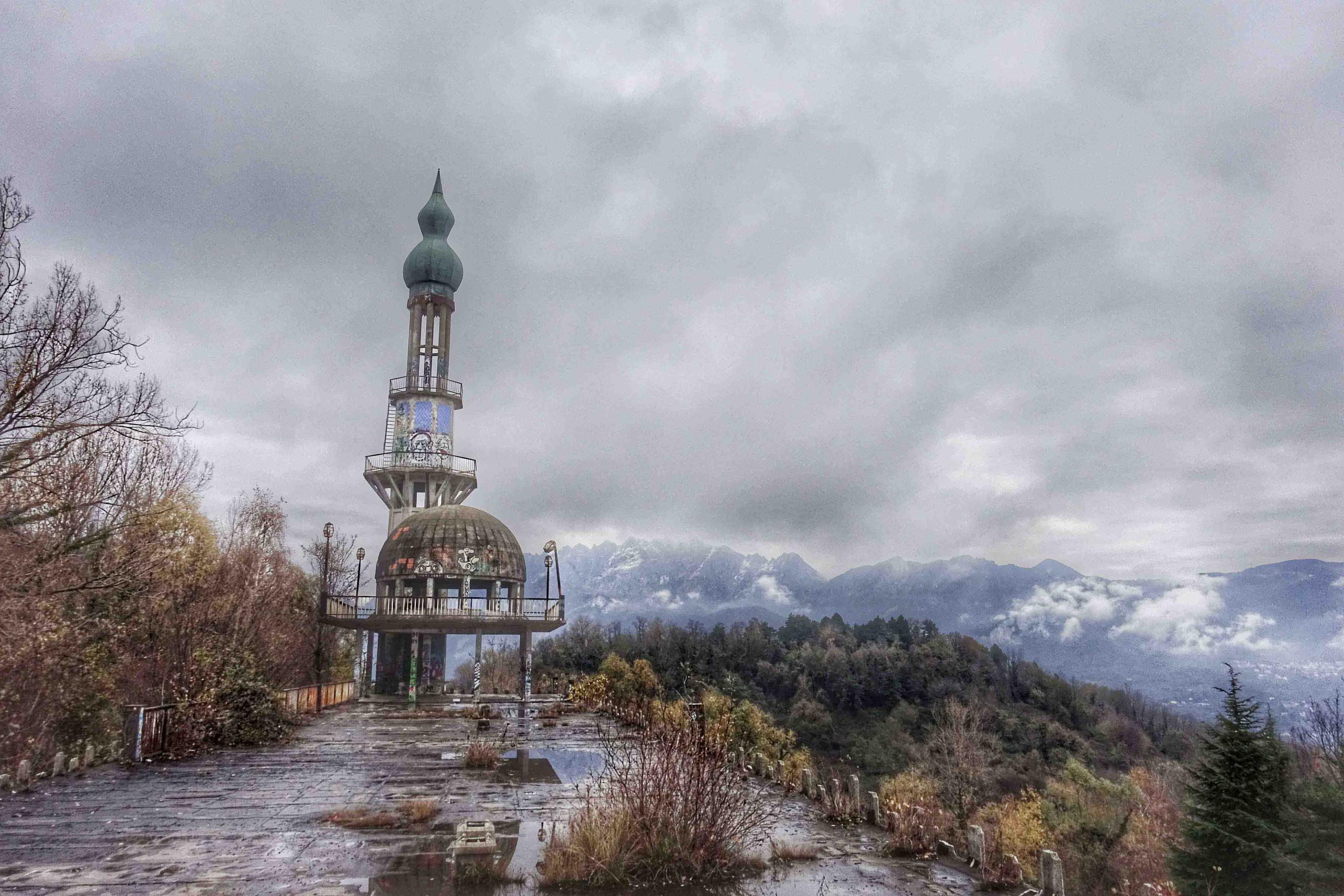 Edificio a forma di minareto visibilmente abbandonato in ambiente boschivo trasandato; cielo nuvoloso