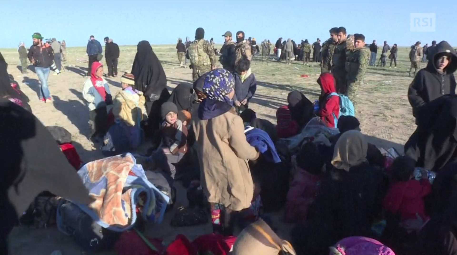 Vista di persone sparpagliate in un campo profughi; in primo piano famiglie con piccoli; in lontananza militari