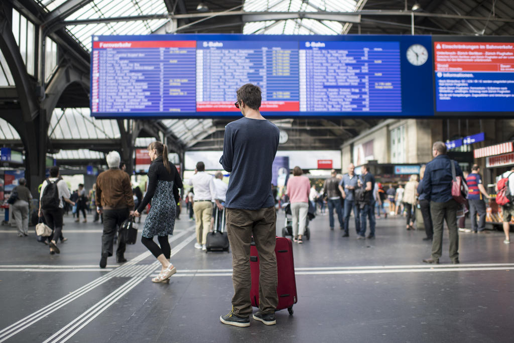 Viaggiatore con trolley fermo in una stazione, guarda verso un tabellone di orari, sfocato, in fondo