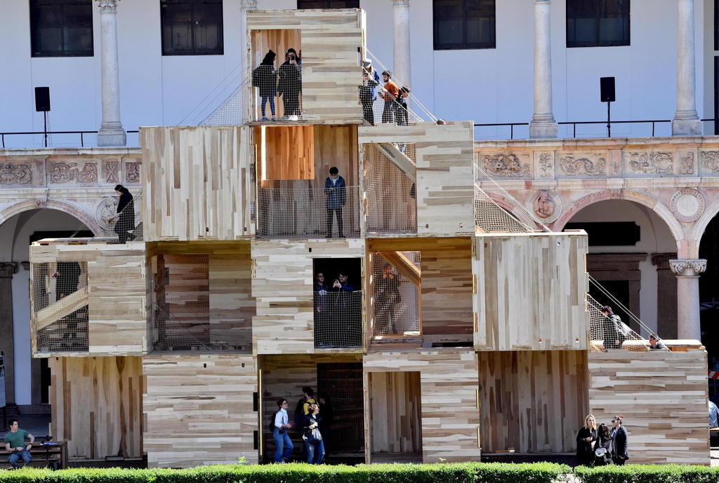 Serie di cubi in legno disposti a piramide su un prato, persone in piedi negli incavi dell installazione.