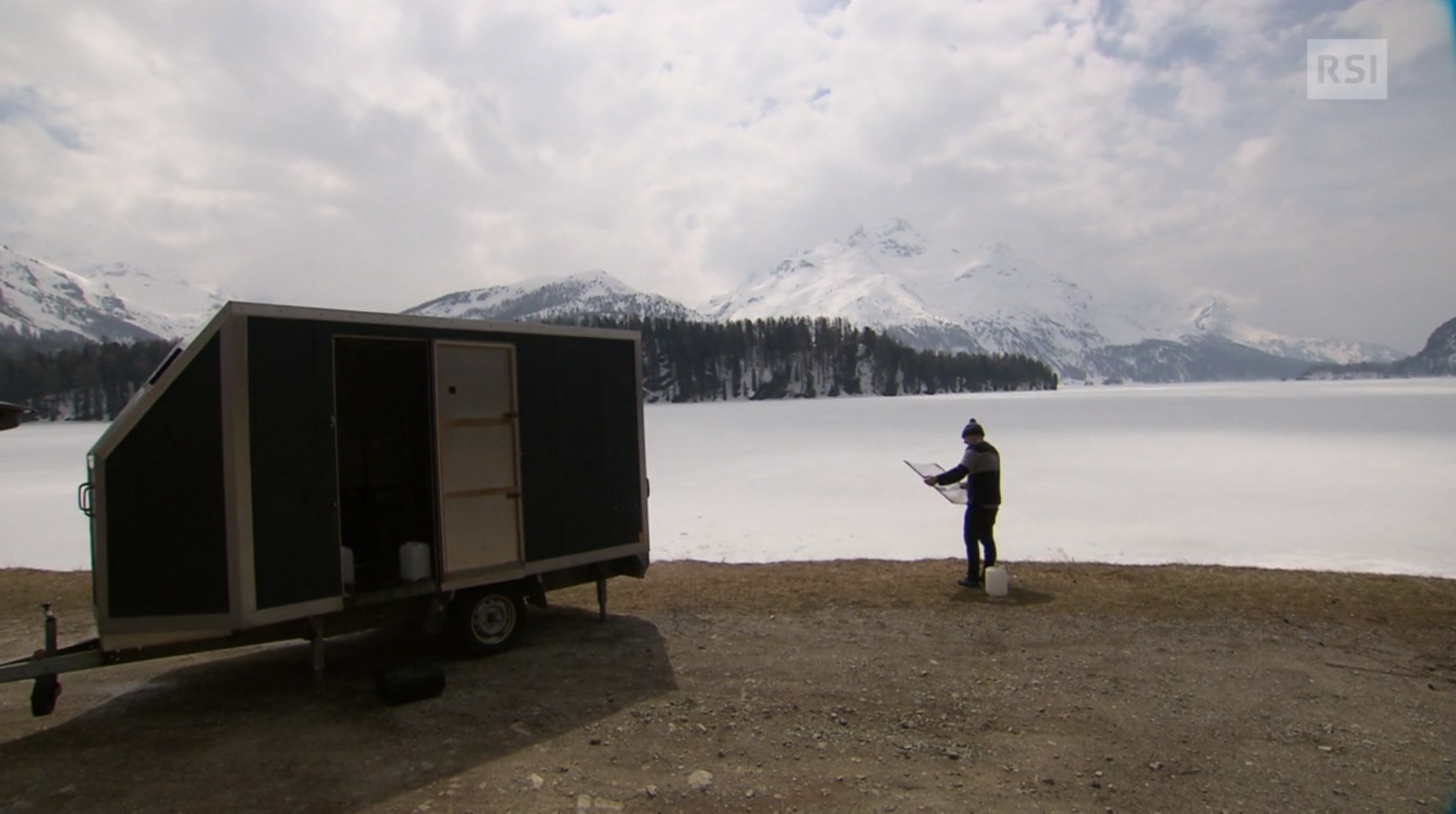 Vista di un lago di montagna; a riva, un rimorchio parcheggiato e un uomo con un telaio in mano