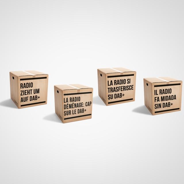 Quattro scatoloni da trasloco con la scritta La radio si trasferisce su DAB+ in tedesco, francese, italiano e romancio