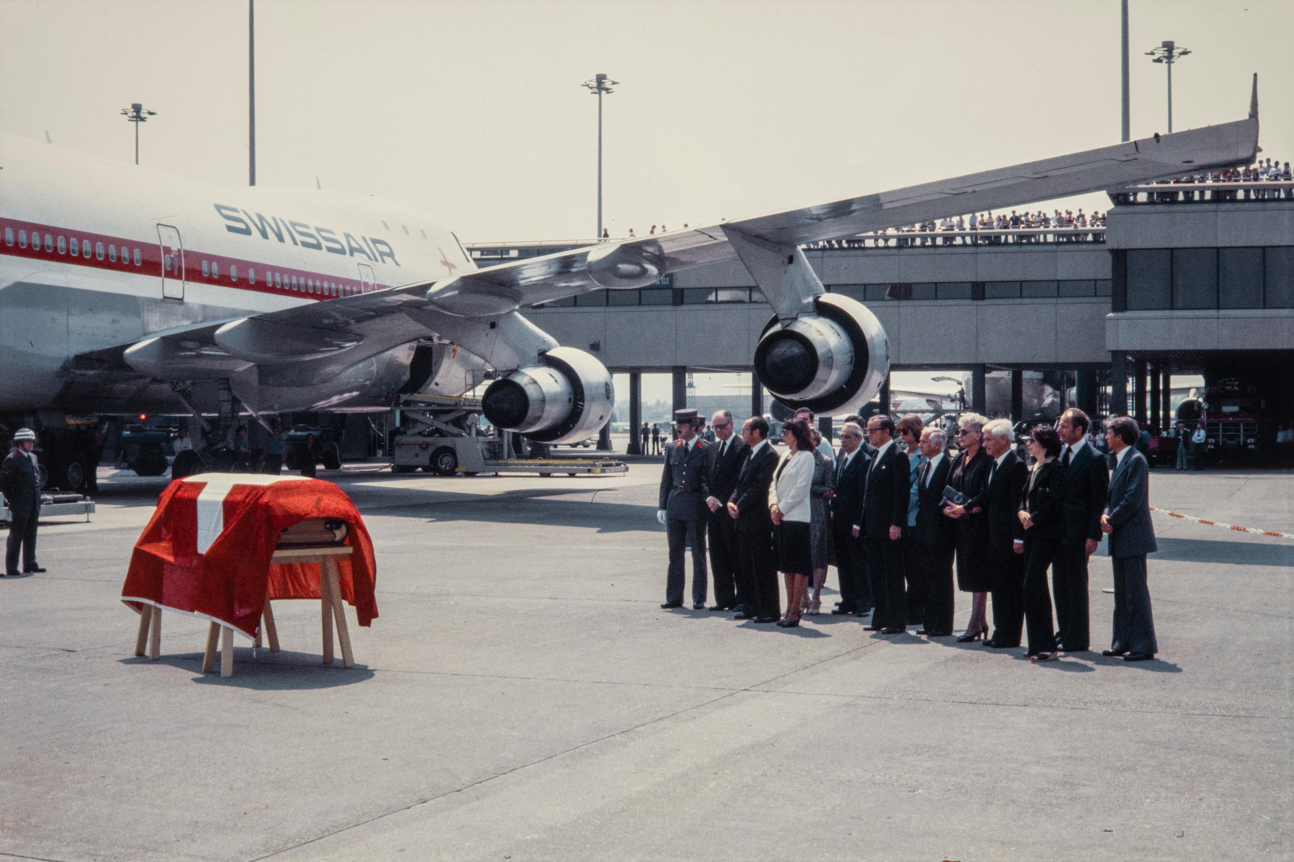 Un féretro con la bandera suiza encima, un avión y un grupo de personas