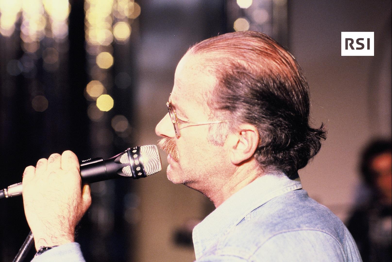 Primo piano di Gino Paoli ripreso di profilo, canta vicinissimo un microfono, dietro luci di scena e uno spettatore (sfocati)
