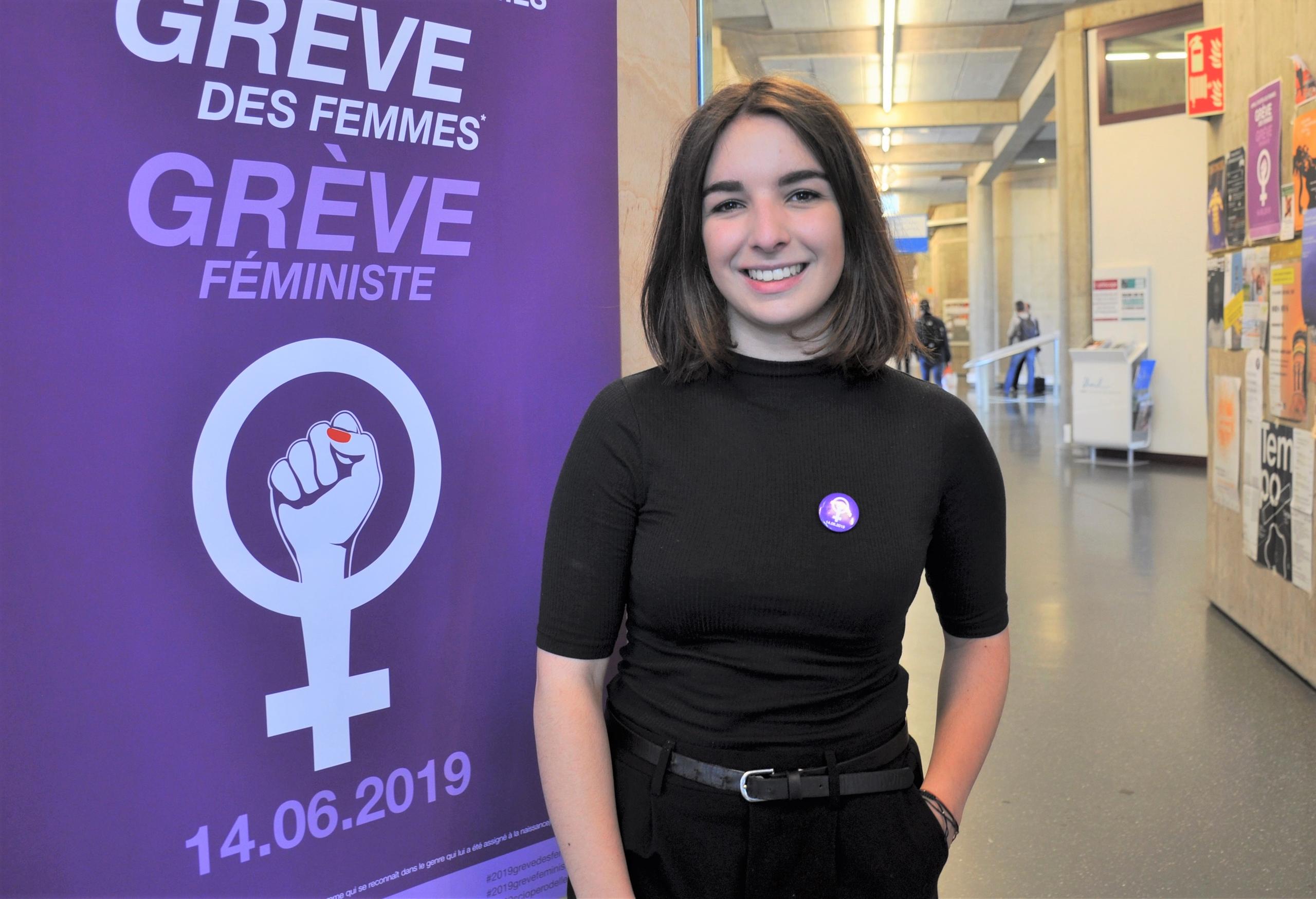 Une jeune femme habillée en noir pose à côté d une affiche violette grève des femmes