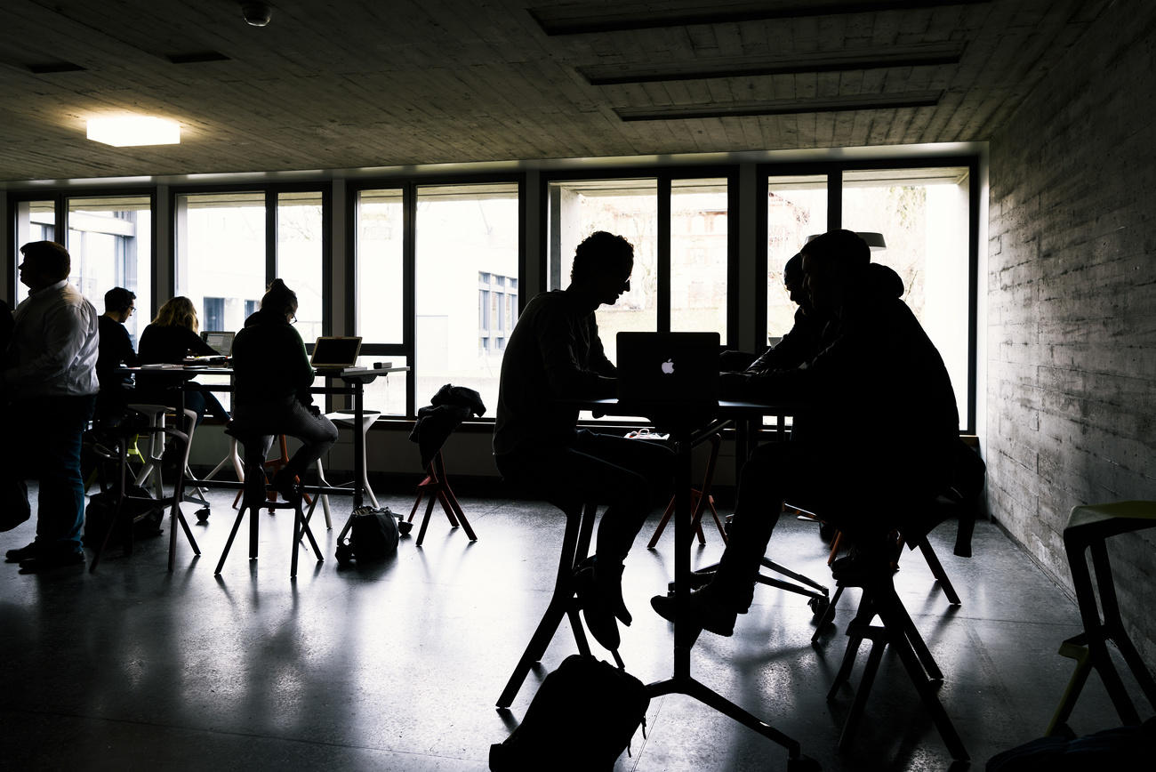 Junge Leute im Gegenlicht aufgenommen sitzten an runden Tischen und arbeiten an Laptops.