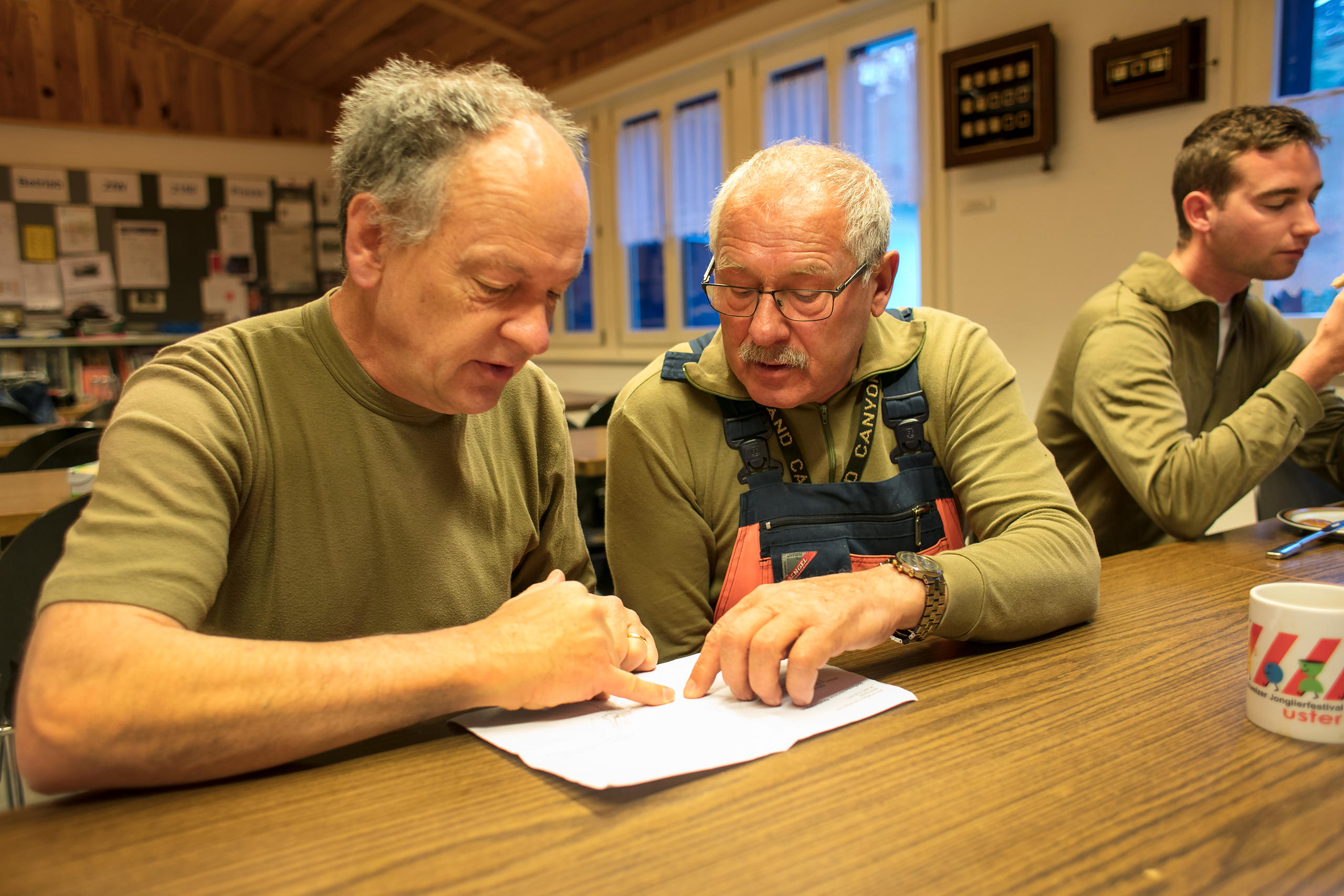 Zwei ältere Männer in feldgrünen Oberkleidern diskutieren an einem Tisch