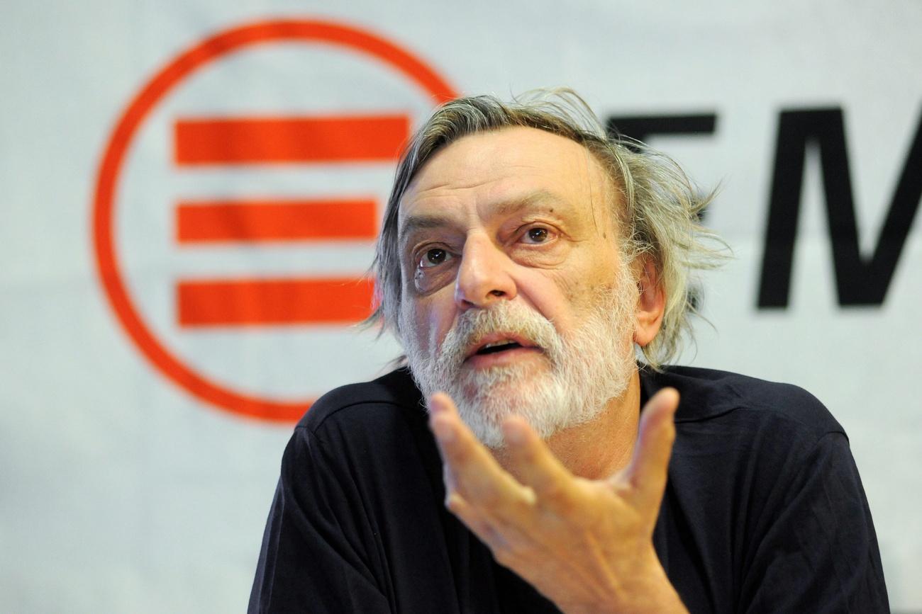 Primo piano di un uomo con capelli e barba bianca; dietro di lui un logo e l inizio della scritta Emergency