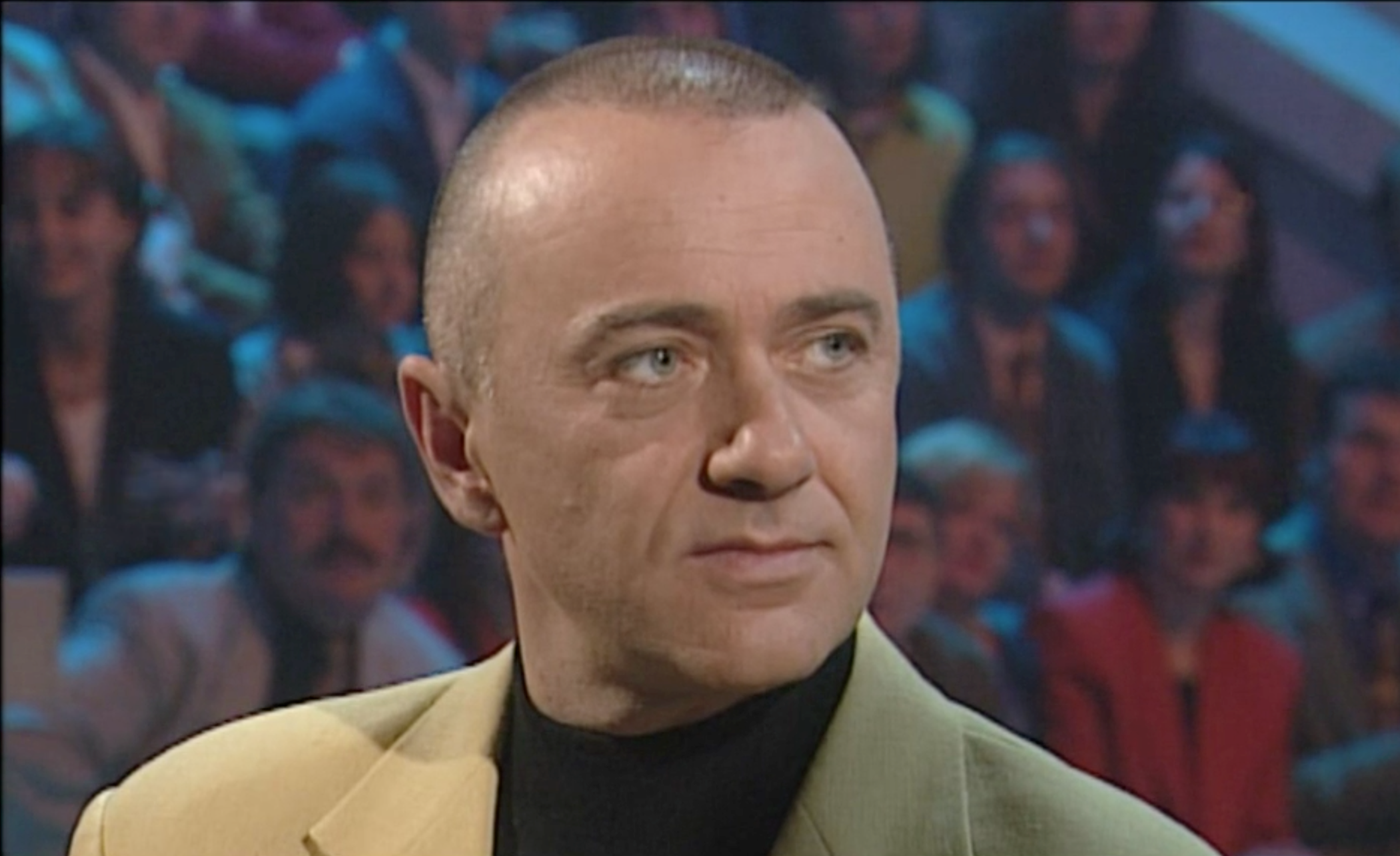 Primissimo piano di Falett con blazer giallo e dolcevita nero; accenna un sorriso; dietro di lui, pubblico