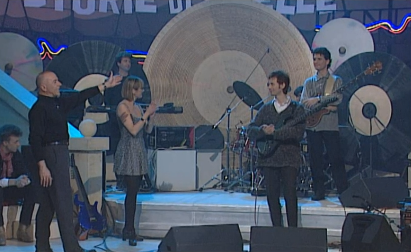 Palcoscenico con un grosso disco di vinile come scenografia; si distinguono un bassista, un chitarrista a una cantante