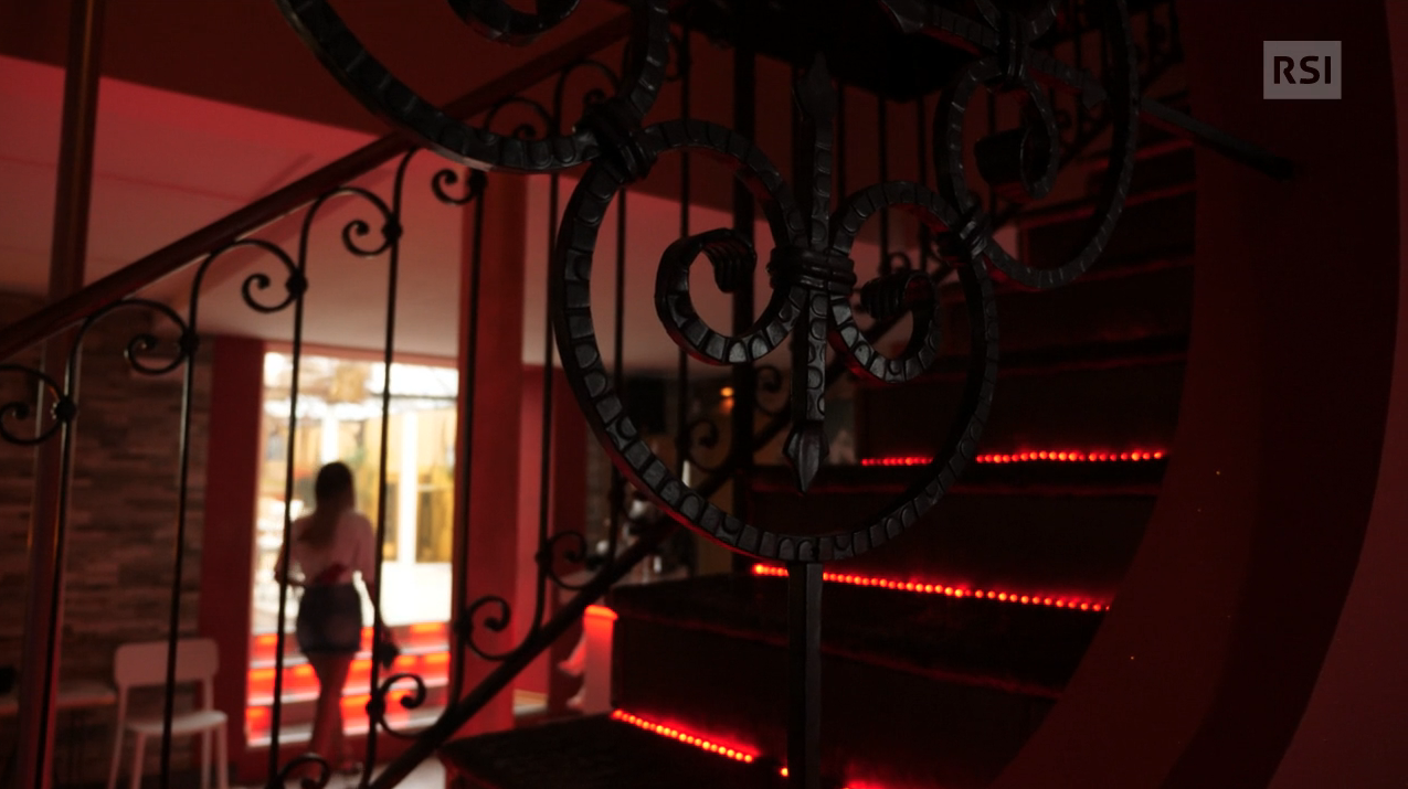 Una donna vista di schiena in un locale in penombra con luci rosse sulle scale in primo piano