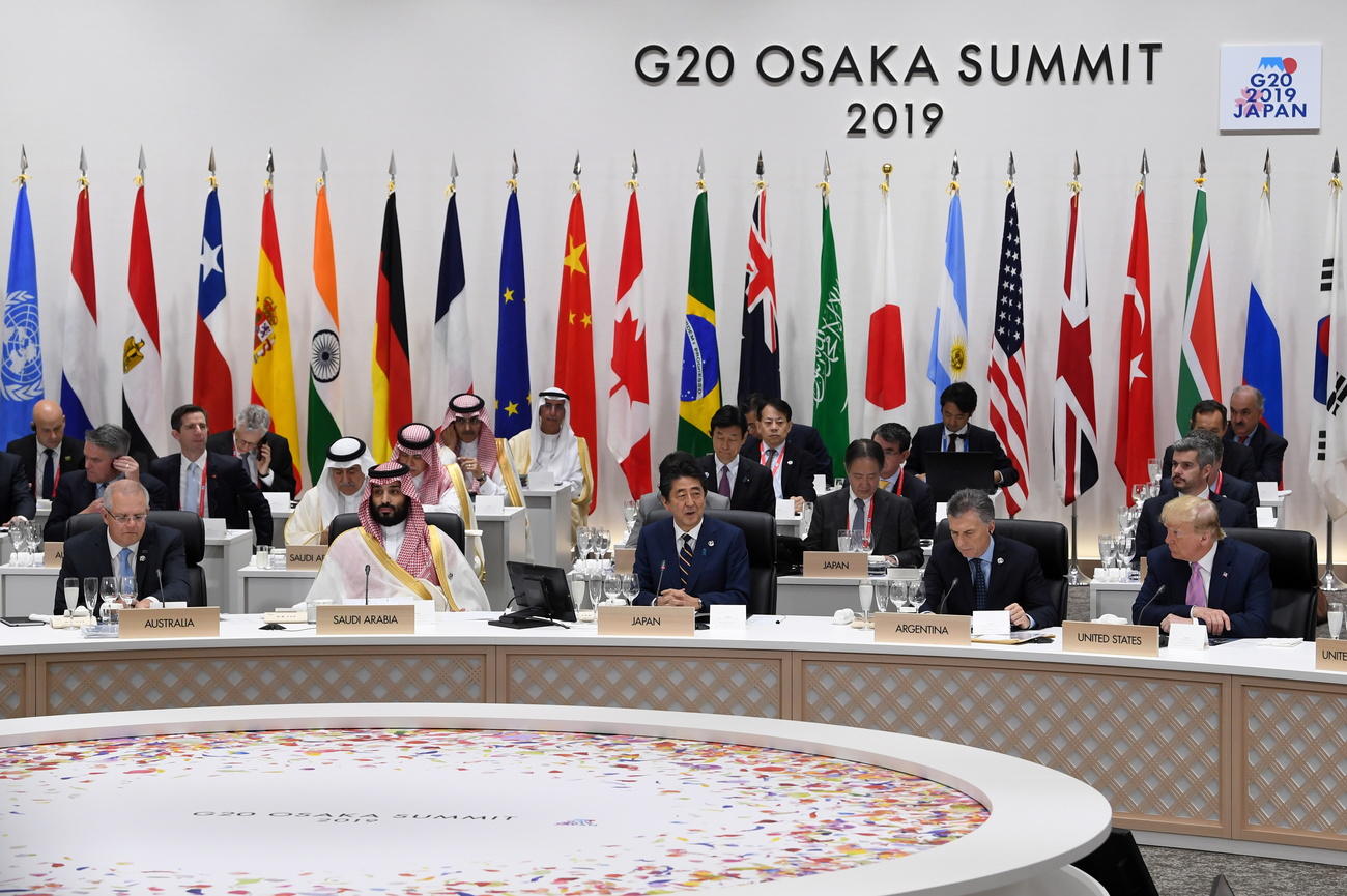Ampio banco leggermente curvo con Abe al centro e altri politici tutt attorno; sul fondo bandiere e logo del G20