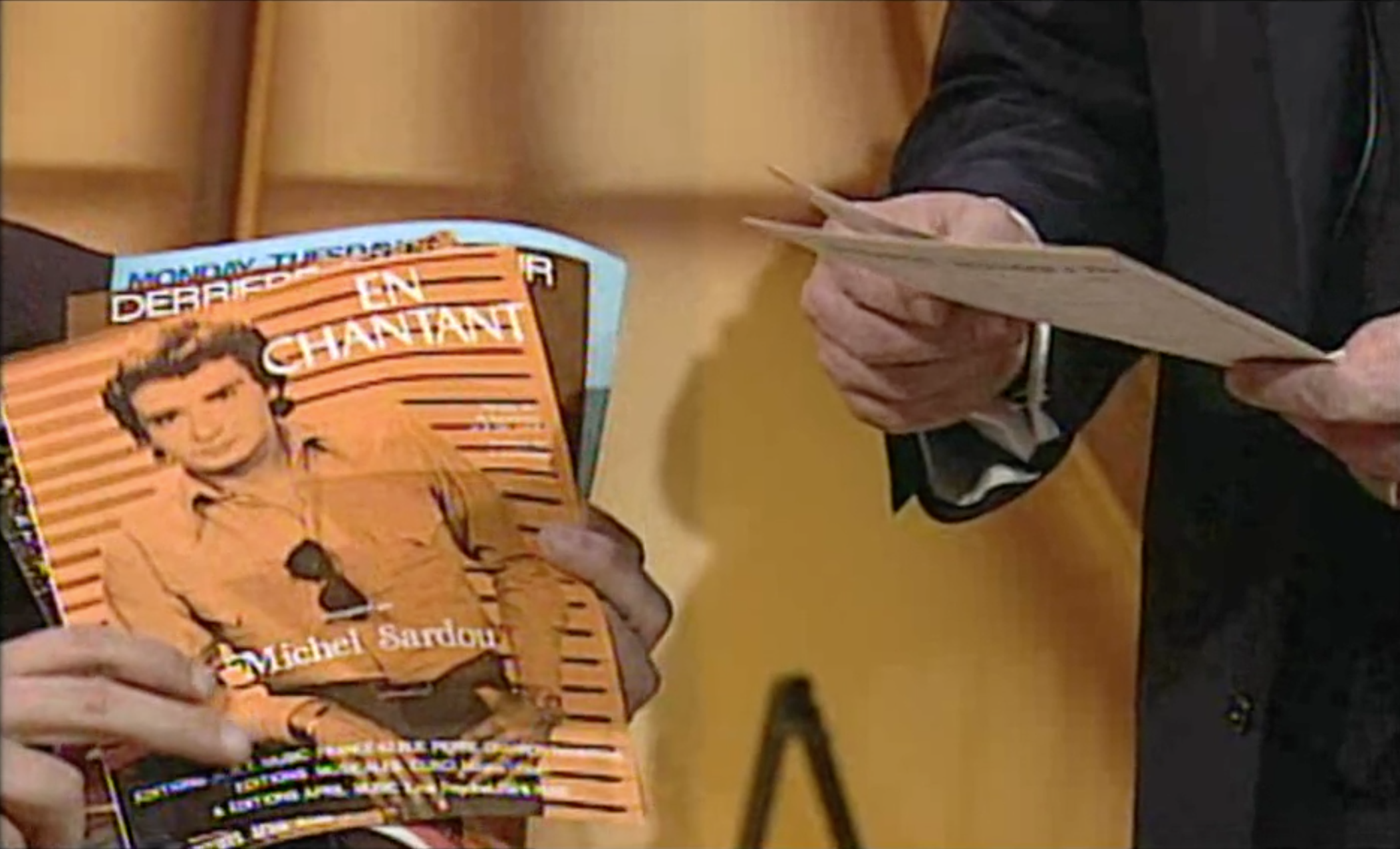 Mani mostrano alla telecamera una risma di fascicoli contenenti spartiti; il primo in alto è En chantant di Michel Sardou