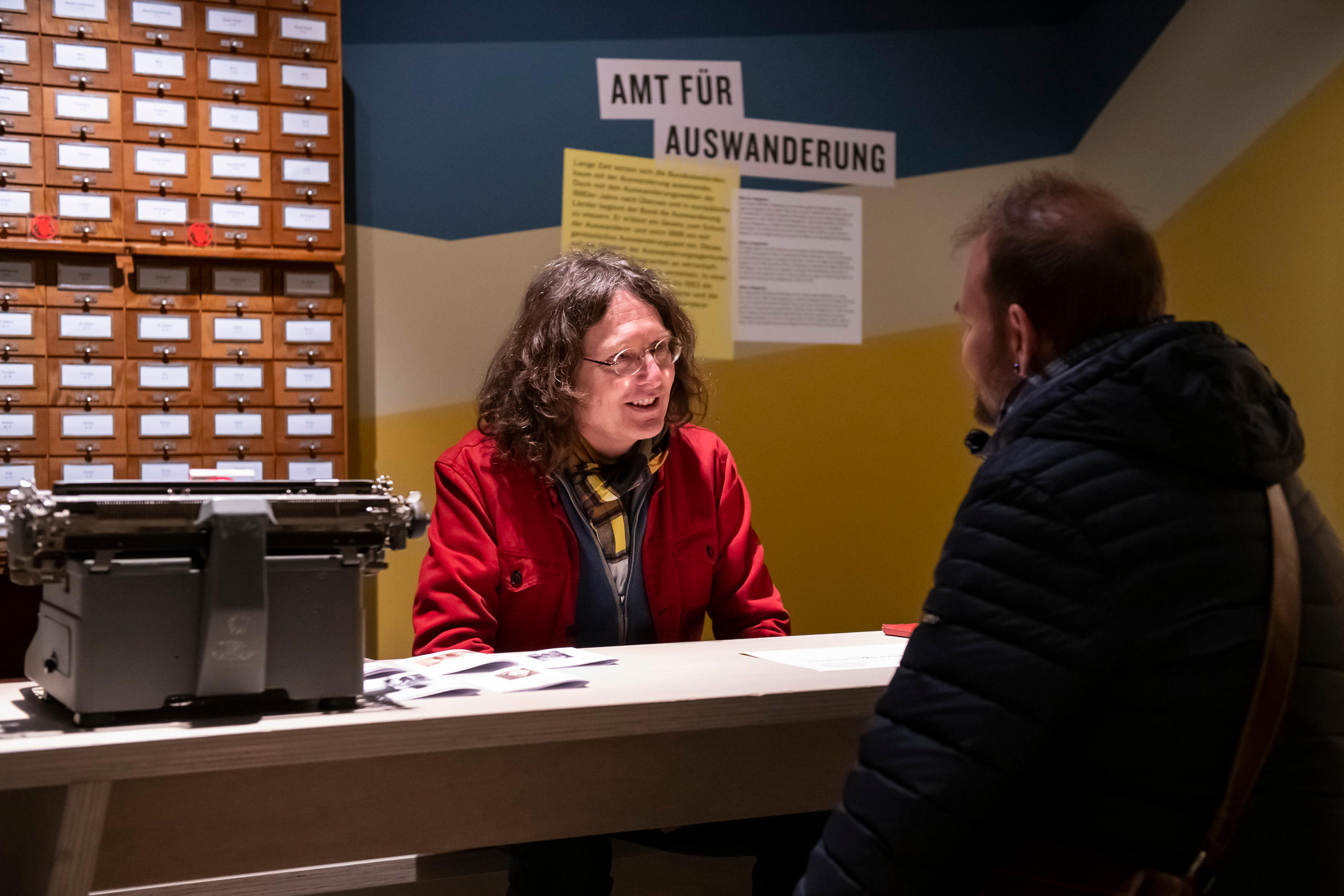Mann hinter und Mann vor dem fiktiven Schalter Amt für Auswanderung an einer Ausstellung