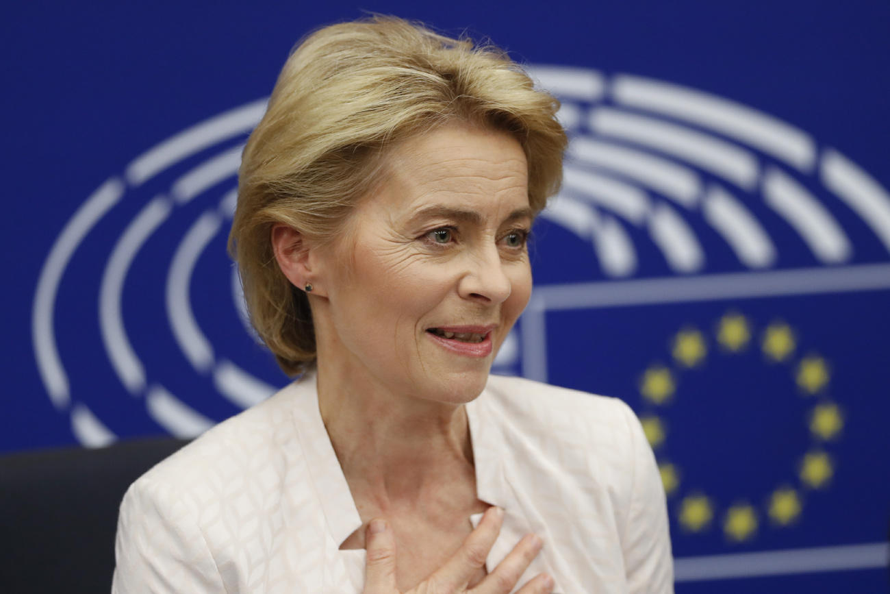 Primo piano di Ursula von der Leyen con, sul fondo, bandiera europea e stemma dell Europarlamento.