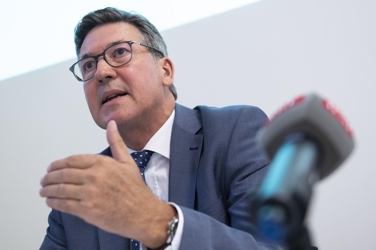 Primo piano di un uomo in abito grigio-azzurro che parla davanti a un microfono gesticolando; nessuno sfondo
