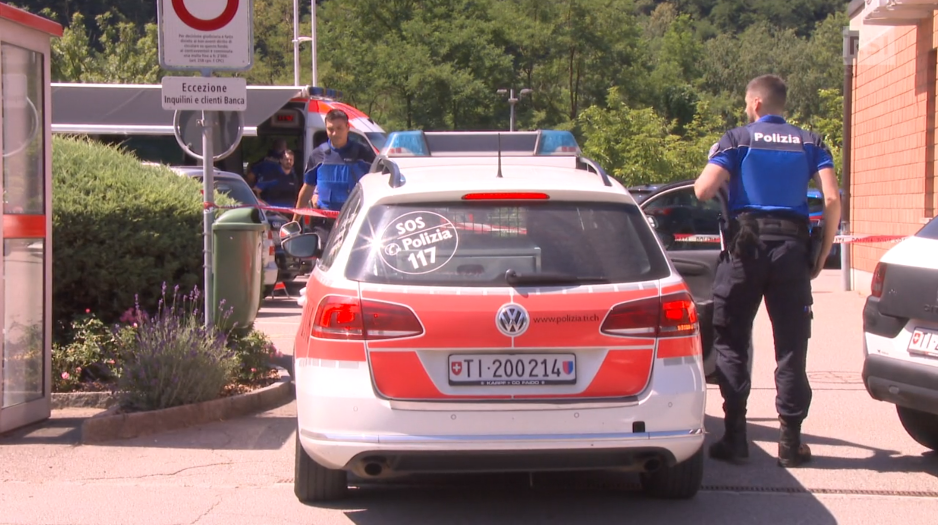 Due agenti -uno di fronte, l altro di schiena- accanto a un auto della polizia; cartello indica la presenza di una banca