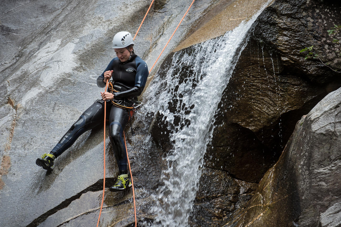A woman wearing helmet and wetsuit abseils down a rock beside a waterfall