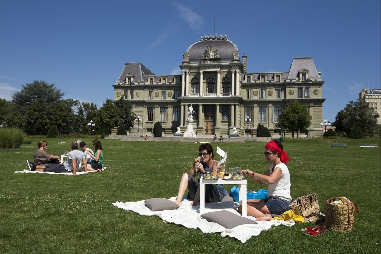 People picnic on the grass. In the background a palatial building.