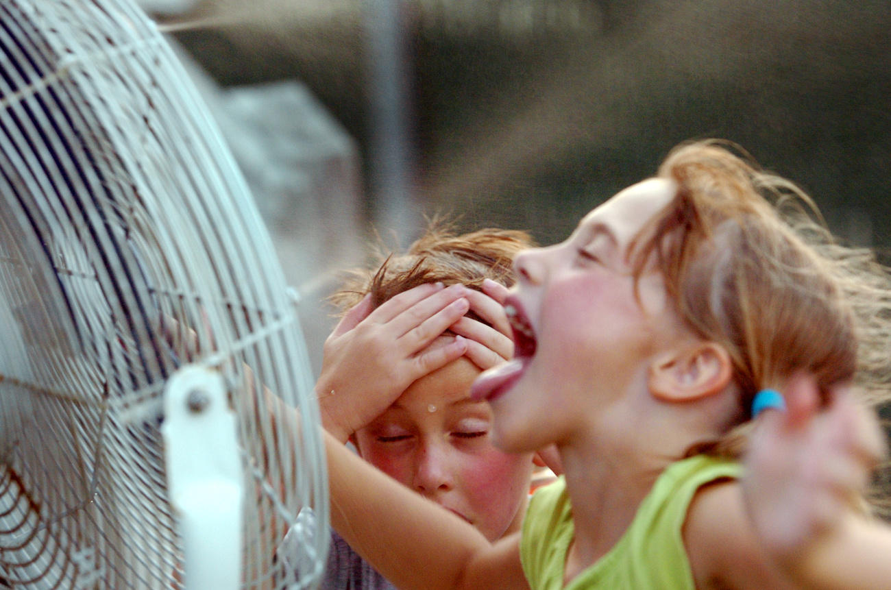 Two children standing in front of a fan seeking relief from the heat
