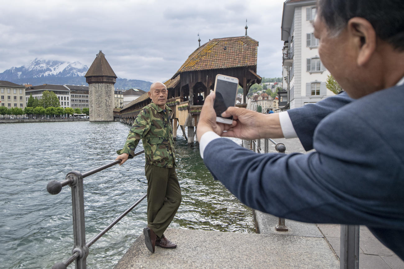 Asian man holds his camera in two hands to take a picture of another man posing in front of Chapel Bridge.
