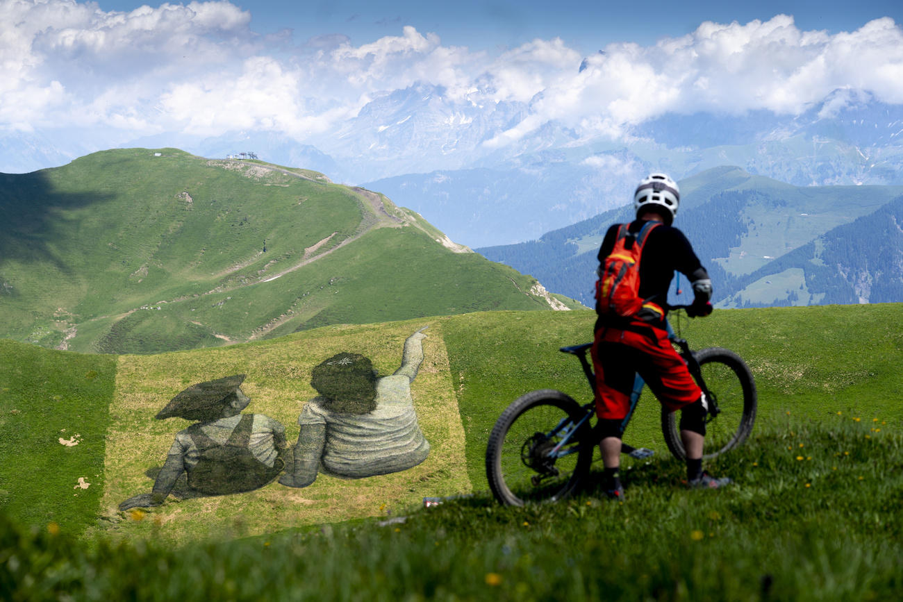 一位山地车骑手正驻足欣赏由法国-瑞士双国籍艺术家Saype在山坡处绘制的可生物降解的巨幅地面涂鸦作品，该作品题为《友谊走私者》。