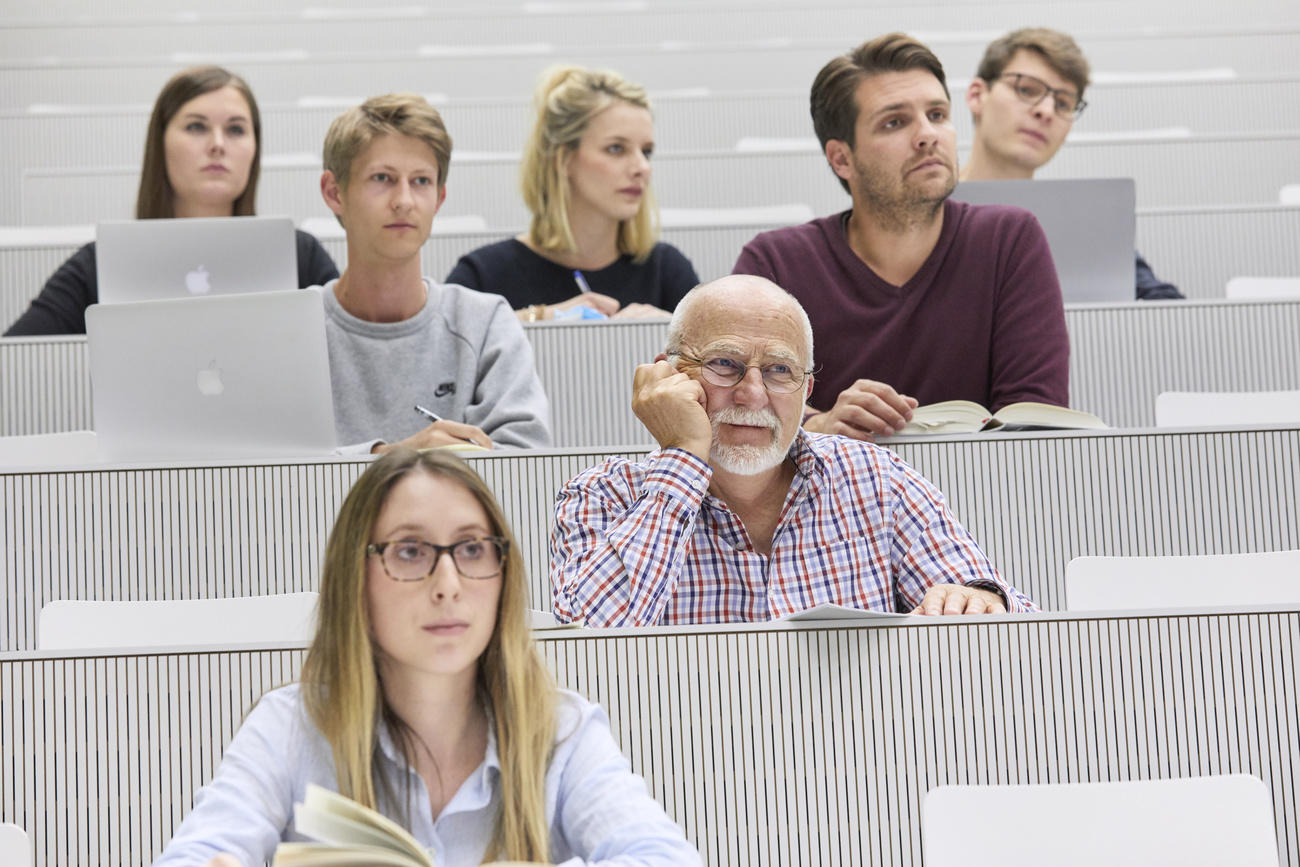 Sette studenti seduti in un auditorio universitario: tutti giovani, a parte un anziano.