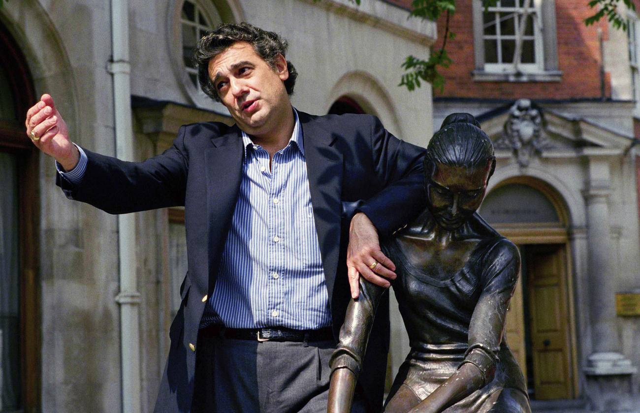 Placido Domingo in camicia e giacca blu posa per i fotografi accanto a una scultura con un espressione ironica