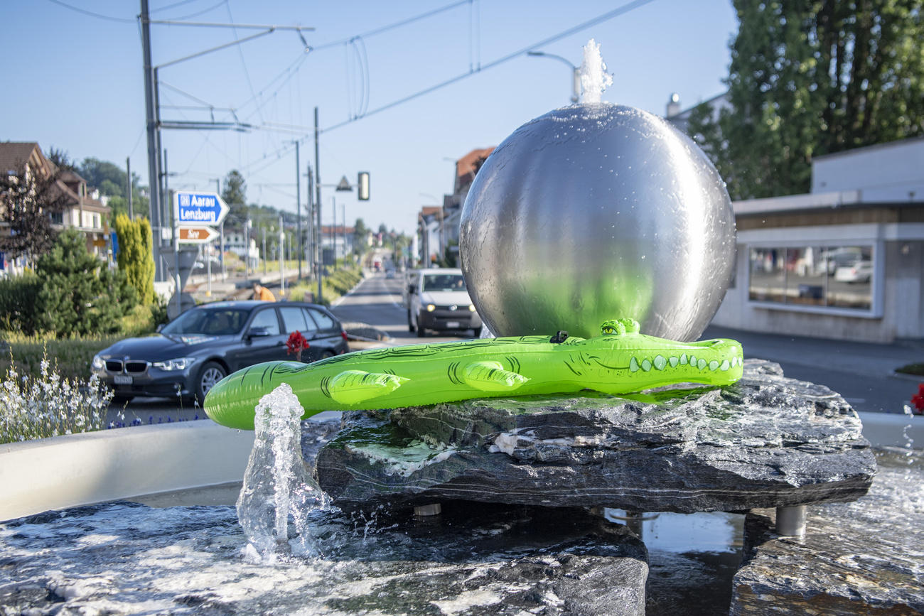 Una fontana cittadina (attorno, strade con auto e cartelli) sulla quale è posato un coccodrillo gonfiabile di plastica