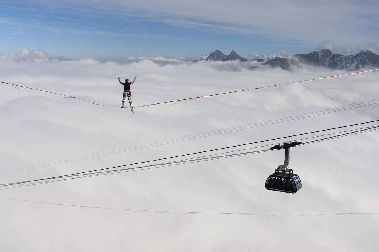 Man on high wire above clouds, with mountaintops in background as well as gondola