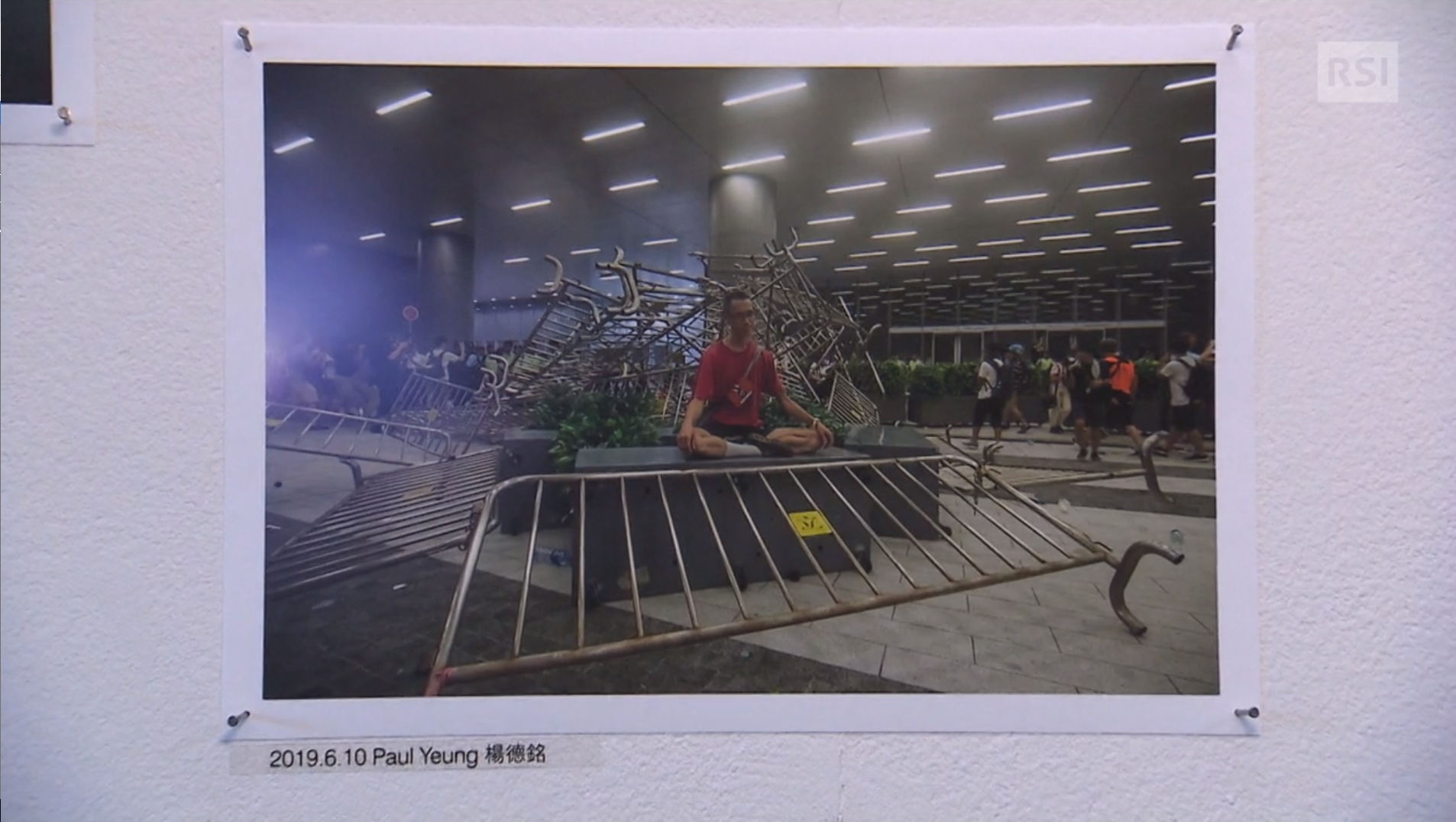 Fotografia appesa a un muro con 4 puntine; mostra un giovane seduto a gambe incrociate in un ambiente caotico