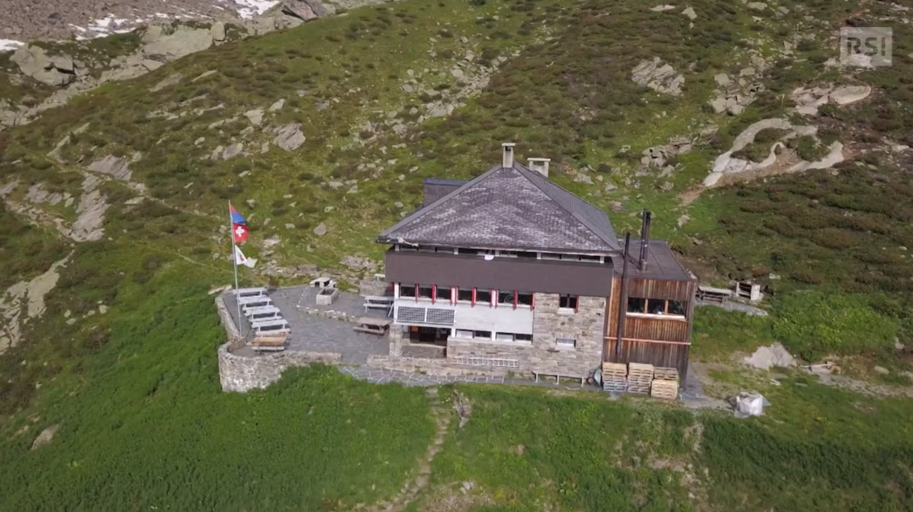 Vista aerea di una costruzione in pietra e legno in paesaggio alpino; accanto a essa, terrazza con bandiere Ticino e Svizzera