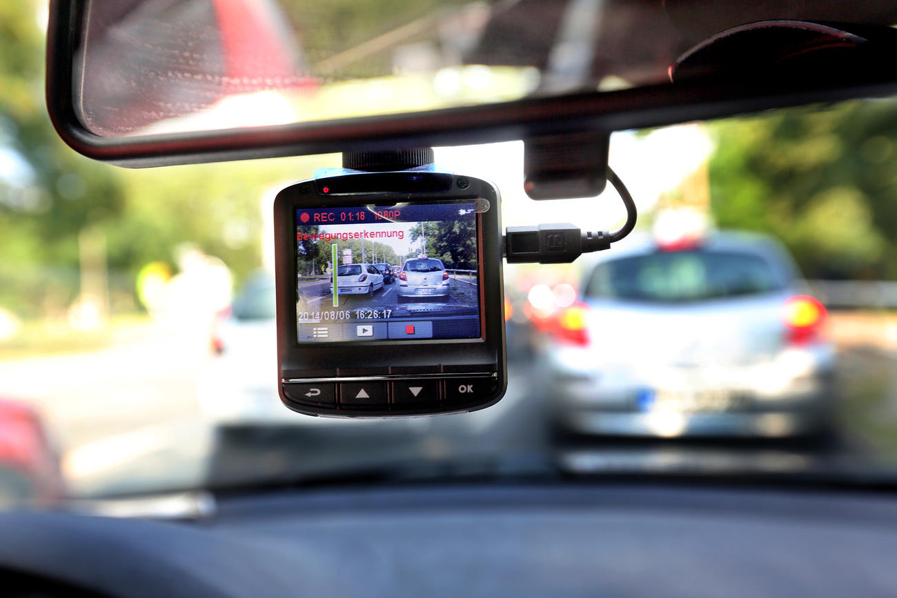 Primo piano di una piccola telecamera applicata a uno specchietto retrovisore di un auto; si intravvedono strada e traffico