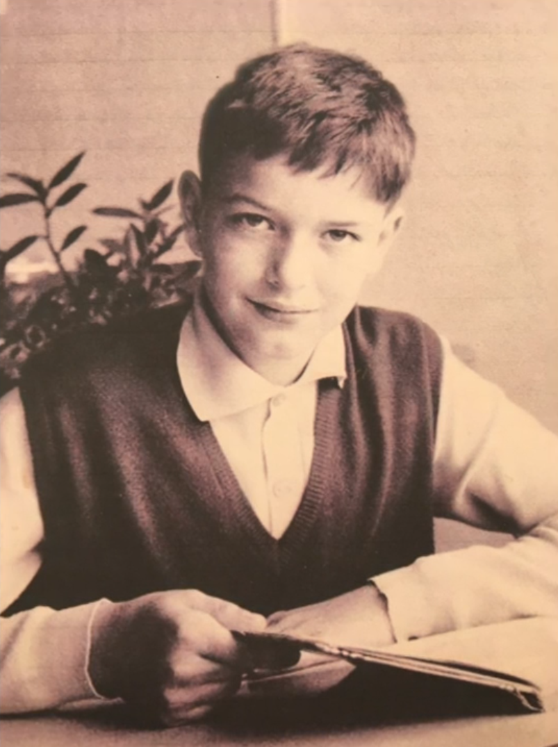 Immagine in bianco e nero di un bambino che, sfogliando un quaderno seduto a un tavolo, sorride alla camera
