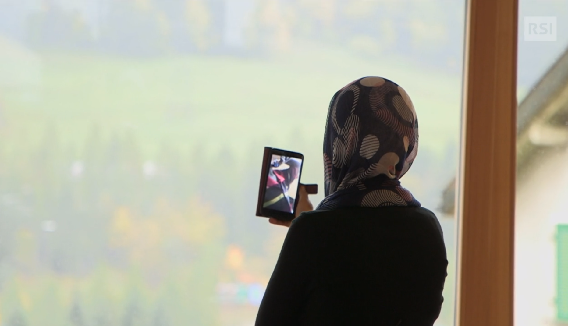 Donna di schiena con velo, rivolta verso una vetrata che dà su un giardino, effettua una videochiamata con lo smartphone