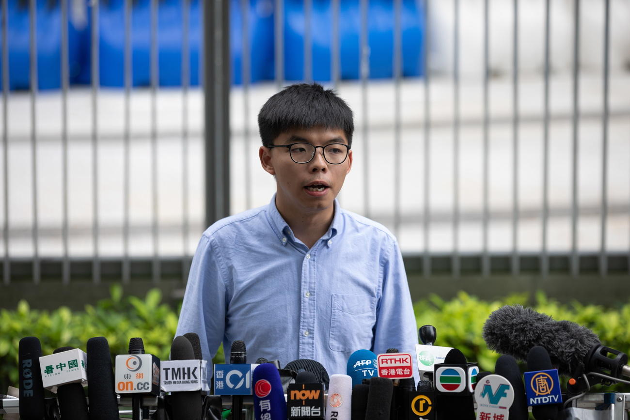 Joshua Wong ritratto a mezzo busto davanti a una decina di microfoni con i loghi delle tv; sullo sfondo, un palazzo