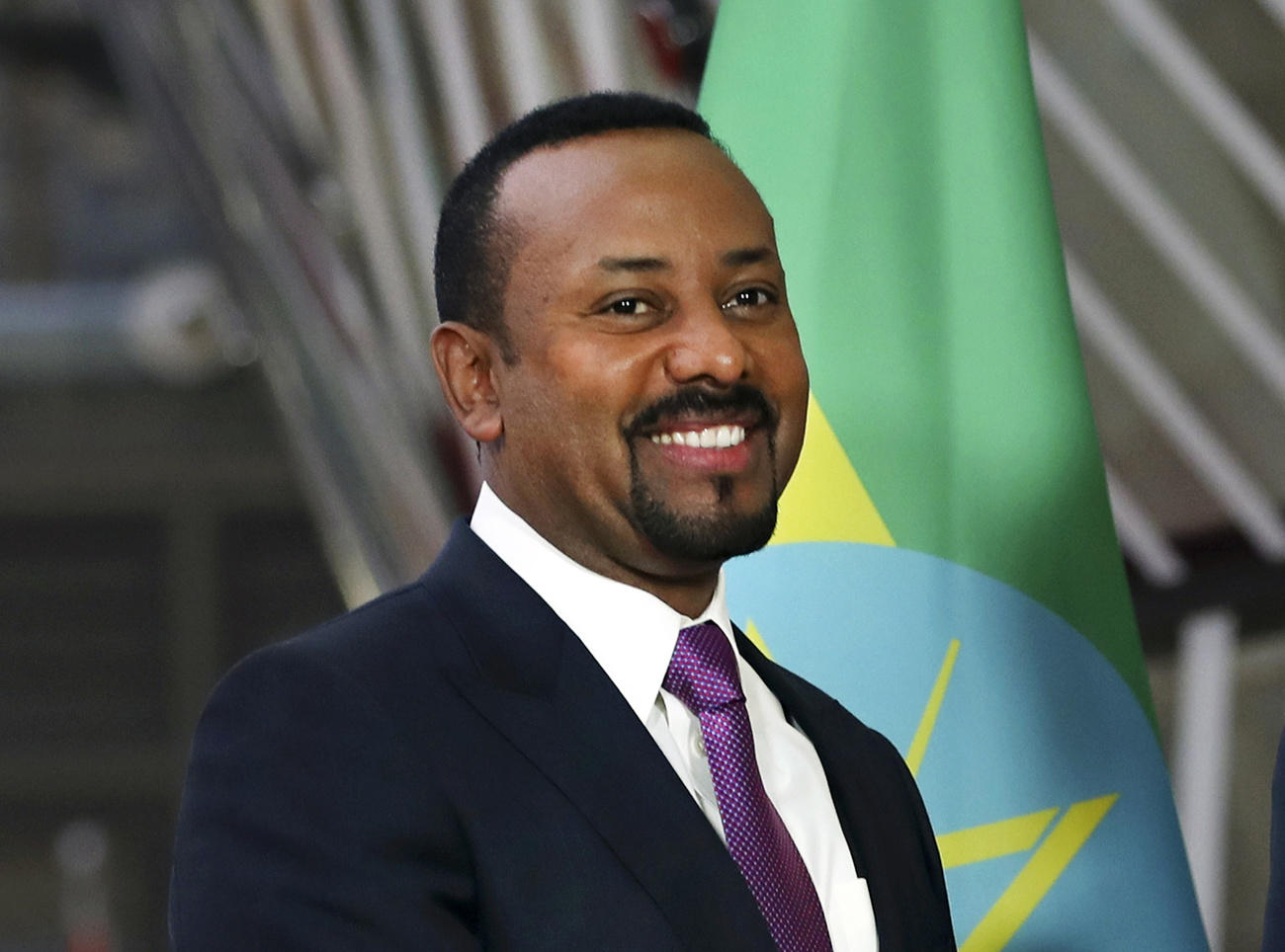 Primo piano di Aby Ahmed Ali, sorridente, con abito scuro e cravatta viola; sullo sfondo, sfocata, bandiera etiope