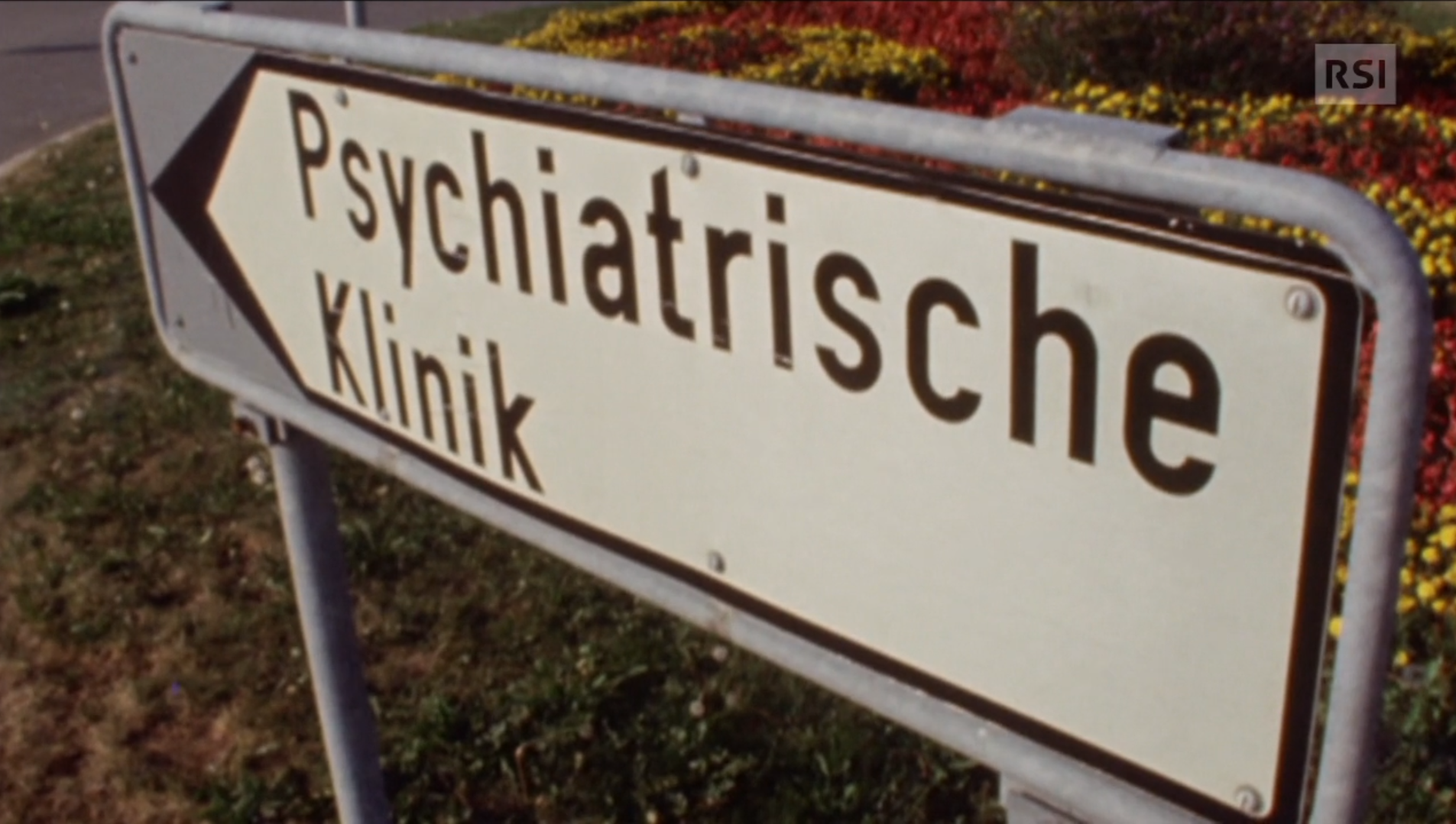 Primissimo piano di un cartello con scritto Psychiatrische Klinik, installato in una aiuola coltivata a fiori