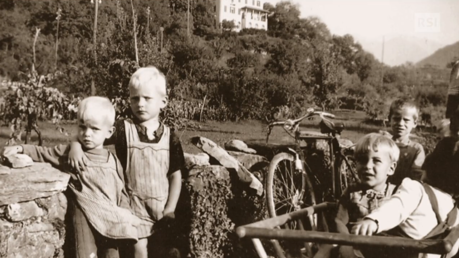 Foto in bianco e nero di quattro bambini con grembiulino vicini a un muretto in paesaggio di campagna