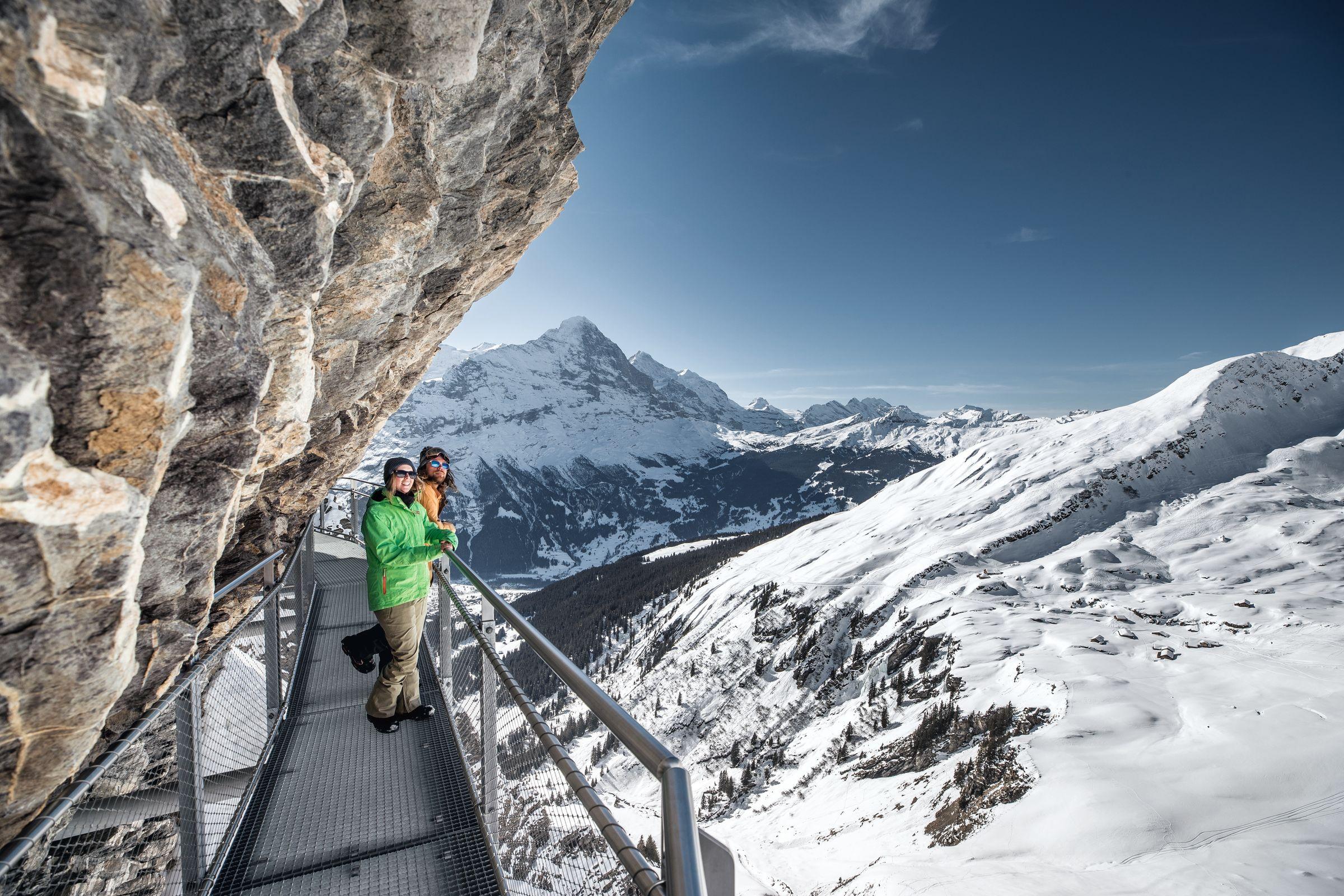 Two people stand on walkway, below cliff, looking at mountain scenery