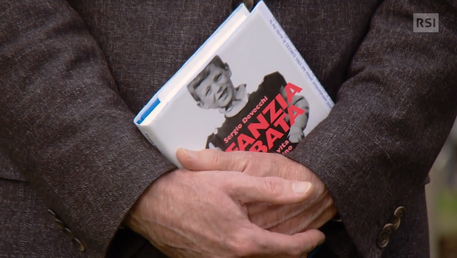 Braccia conserte vestite da giacca scura tengono in mano un libro intitolato Infanzia rubata