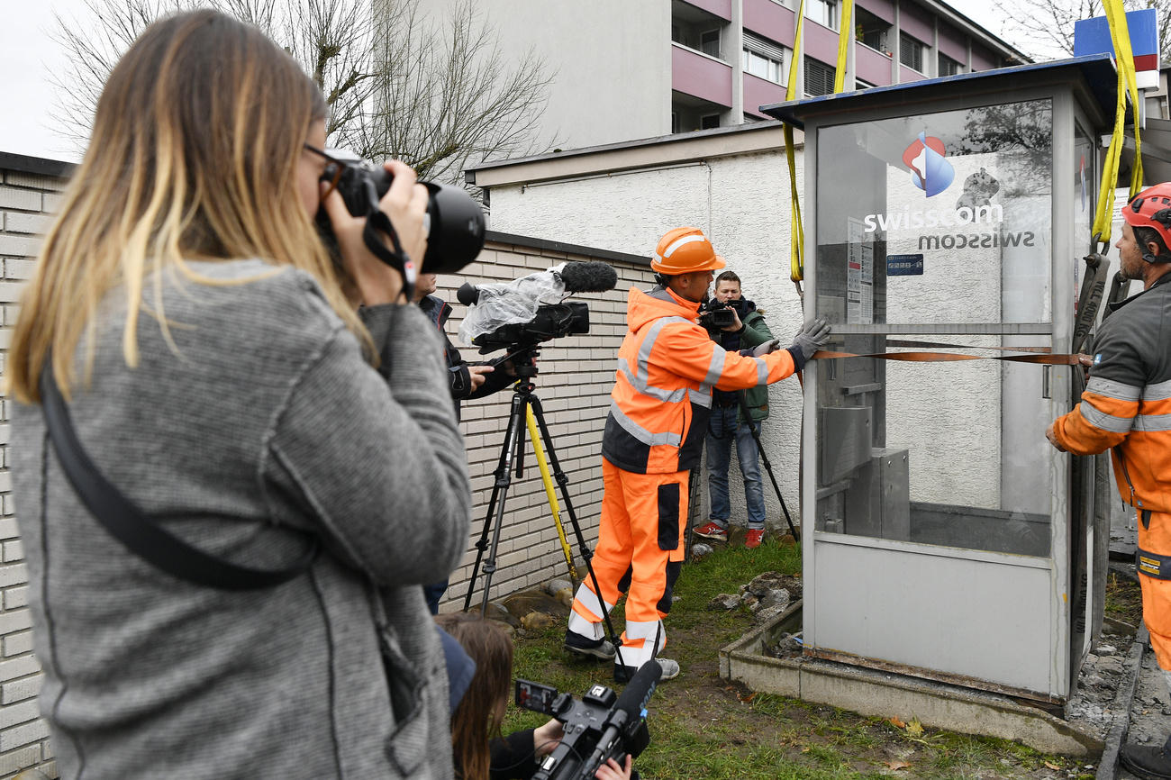 Operai assicurano a un argano una cabina telefonica mentre fotografi e teleoperatori riprendono la scena