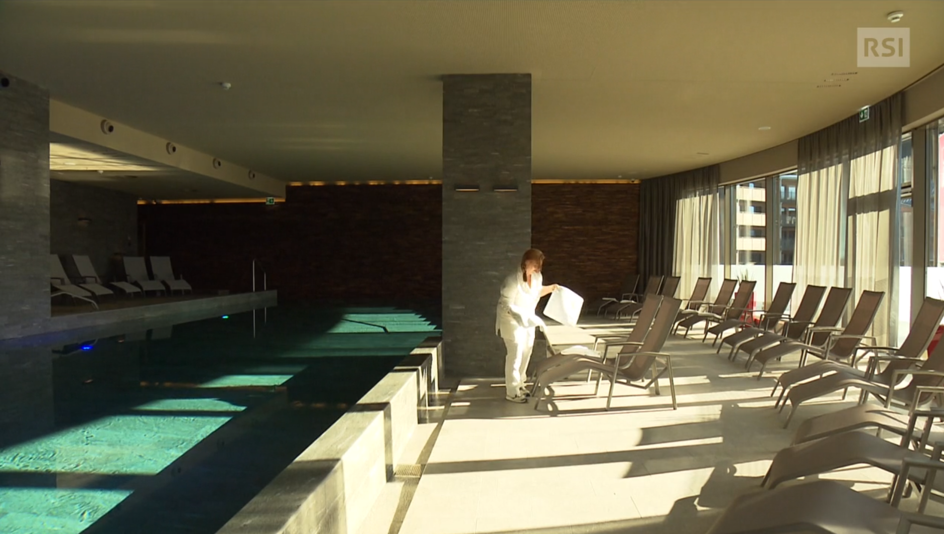 Interno di una piscina coperta; un inserviente sta posando un asciugamano su una chaise longue