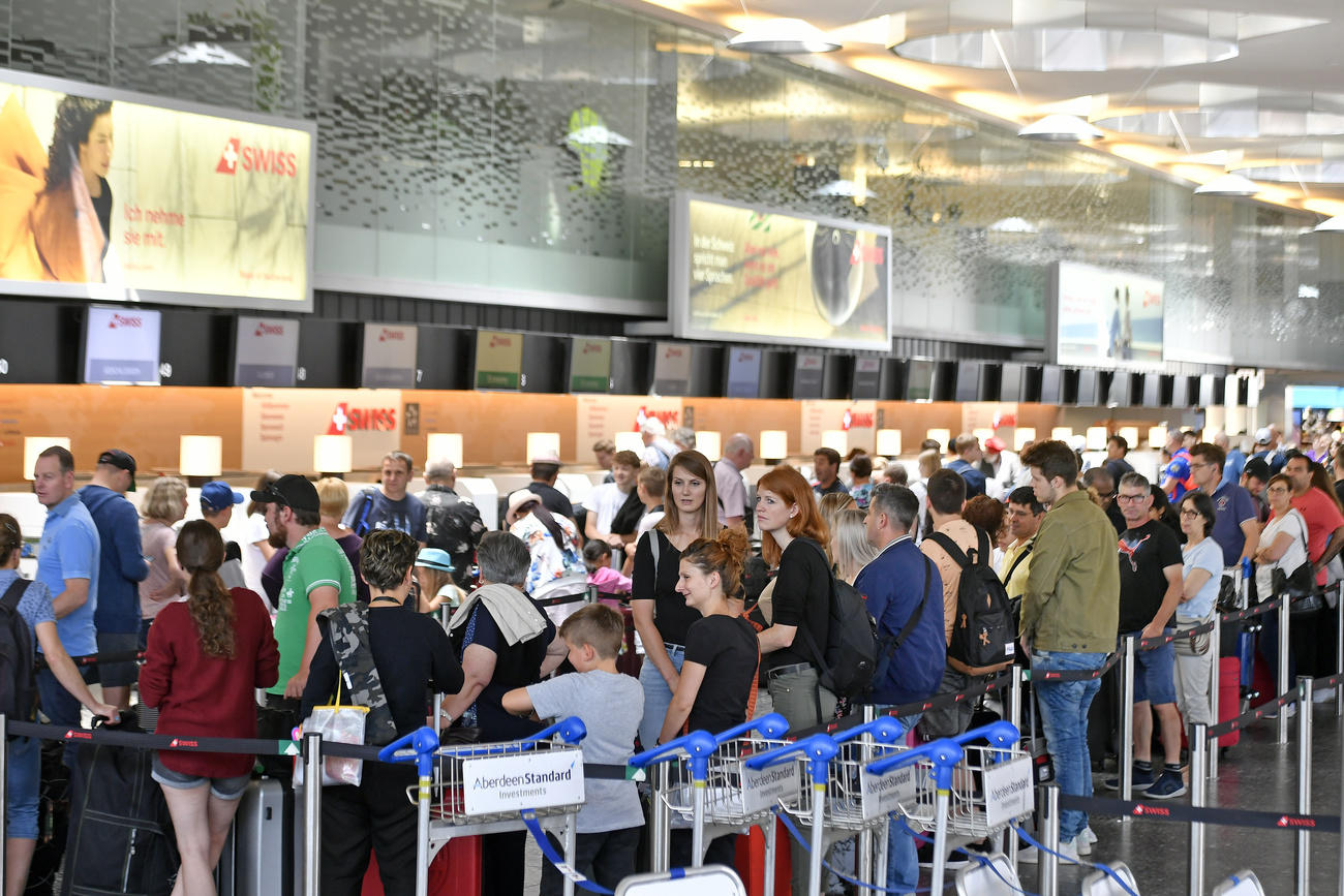 Immagini dei persone con valige e carrelli in coda in una sala di aerostazione; schermi sopra il banco