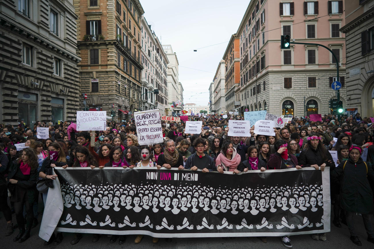 Immagine frontale di corteo tra le strade d una città con palazzi antichi; in testa striscione con ritratto un gruppo di donne