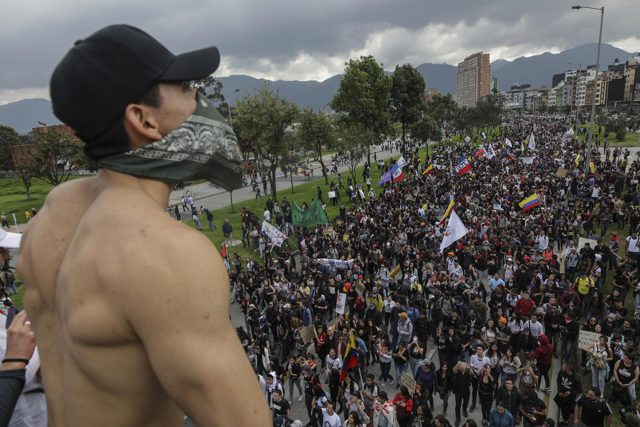Un corteo di protesta di migliaia di persone visto da un punto elevato (forse un terrazzo) dove sosta un uomo con volto coperto